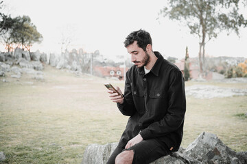 Confident young Hispanic man using a smartphone in the park, wearing a black jacket