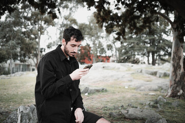 Confident young Hispanic man using a smartphone in the park, wearing a black jacket