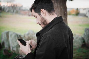 Confident young Hispanic man using a smartphone in the park, wearing a black jacket