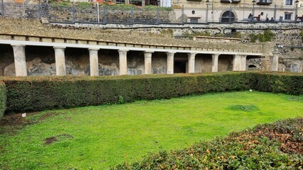 Herculaneum, Italy - 9 January 2025. A grass courtyard with hedge borders stands before a long...