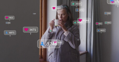 Woman wearing grey bathrobe sipping from white mug by wooden window at home, with social icons