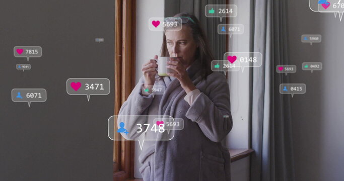 Woman wearing grey bathrobe sipping from white mug by wooden window at home, with social icons