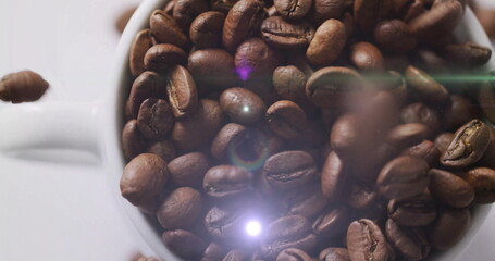 Falling roasted coffee beans filling white ceramic mug on smooth white surface, with lens flare