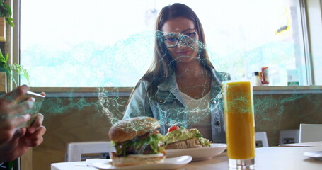 Woman wearing denim jacket, glasses sampling avocado toast, drinking juice, checking phone at cafe