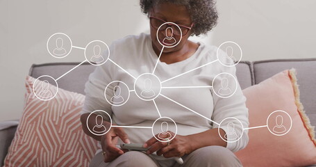 Using smartphone woman with glasses sitting on sofa with peach cushions at home, with network icons