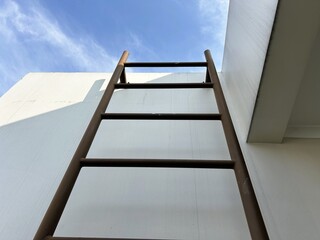 Upward view of a simple metal ladder against a white building wall and blue sky