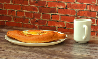 Khachapuri, traditional Georgian pie with baked egg and cheese on a wooden table, next to a cup of hot black tea.