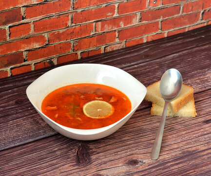 Solyanka, tasty soup in a deep bowl with a slice of lemon on a wooden table, a tablespoon and two pieces of wheat bread nearby.