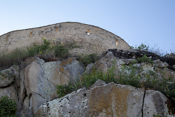 stone fortress wall sits atop a rugged, weathered rock cliff covered in green vegetation and ivy under a clear blue sky. a historical landmark