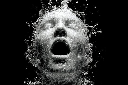 Dramatic black and white image of a human face emerging through splashing water, mouth open in an expressive moment, creating a powerful and surreal visual effect