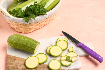 Wicker basket with fresh green zucchini and board of slices on pink background