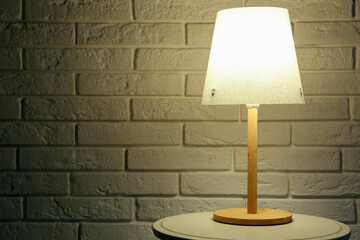 Glowing lamp on wooden table near white brick wall in dark room