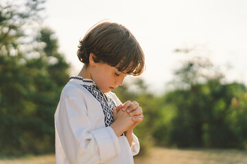 The child stands with hands together in a prayer pose, wearing a traditional embroidered shirt,...