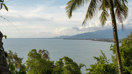Scenic tropical coastline with calm sea, lush greenery, palm tree foreground, and distant mountains under a partly cloudy sky.