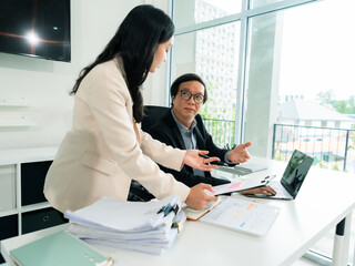 An old businessman and two Asian female secretaries are talking and working together using a laptop and analyzing documents with charts and graphs to solve financial problems in a company.