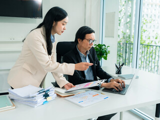 An old businessman and two Asian female secretaries are talking and working together using a laptop and analyzing documents with charts and graphs to solve financial problems in a company.