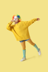 Funny little girl in clown wig with party blower on yellow background. April Fools' Day celebration