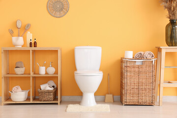 Interior of restroom with toilet bowl, shelving unit and basket near orange wall