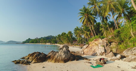 Aerial view of Koh Samui's pristine beach, featuring lush palm trees and rocky shores. The serene turquoise waters and clear blue sky create a tranquil tropical escape