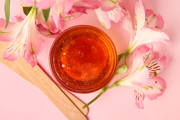 Glass bowl with sugaring paste, wooden sticks and flowers on pink background, closeup