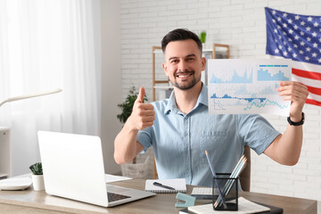 Handsome young happy man with laptop, graphics and flag of USA showing thumb-up gesture in office