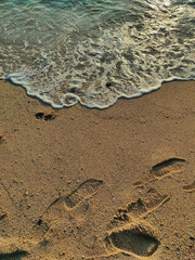 Footprints in the golden sand lead towards the sea, where a gentle, foamy wave glitters in the sunset light.