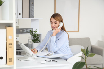 Young businesswoman with document folder talking by telephone at desk in office