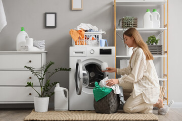 Young woman putting laundry detergent and dirty clothes into washing machine in bathroom