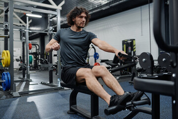 Young Caucasian man with curly hair doing strengthening exercise in modern gym using cable row machine