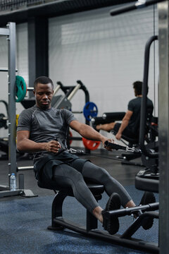 Young African American man having strengthening workout in modern gym using cable row machine, vertical long shot