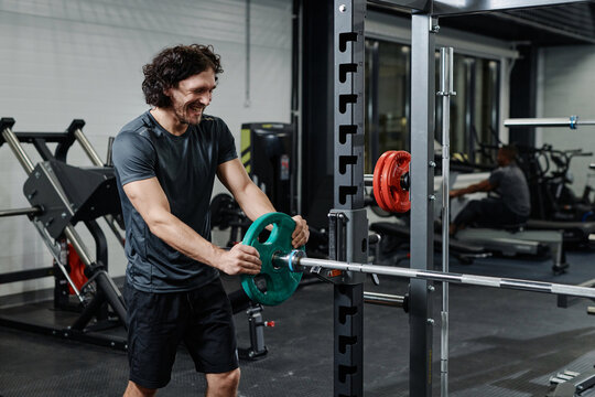 Cheerful young Caucasian man changing weights on barbell for strengthening exercise in modern gym, copy space