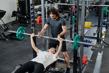 Young Caucasian trainer helping sporty woman with bench press exercise in modern gym, copy space