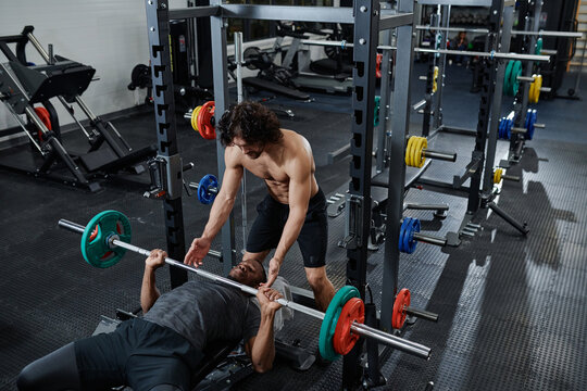 High angle view wide shot of young Caucasian trainer spotting Black man during bench press exercise in gym