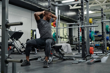 Wide shot of young Black man sitting on bench in gym doing stretching exercise, copy space
