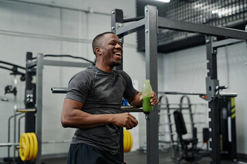 Young Black man holding fresh smoothie drink in bottle laughing while chatting with someone in gym