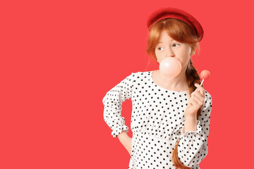 Cute ginger teenage girl with lollipop blowing bubble gum on red background