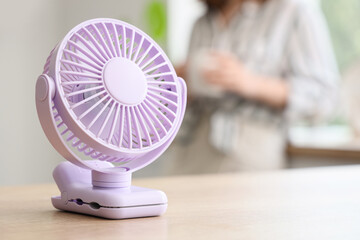 Mini fan on table against woman with cup in kitchen, closeup