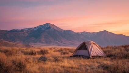 A lone tent sits in a dry, grassy field at sunset, with snow-capped mountains in the background under a soft, pink and purple sky