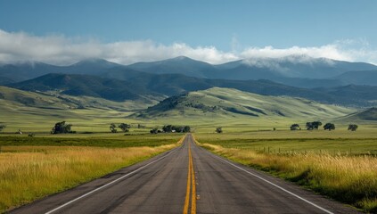 Naklejka premium A long, straight asphalt road vanishes into a valley nestled between majestic, hazy mountains under a clear blue sky
