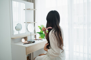Woman brushing long hair at a brightly lit vanity mirror in a cozy room with natural light and...