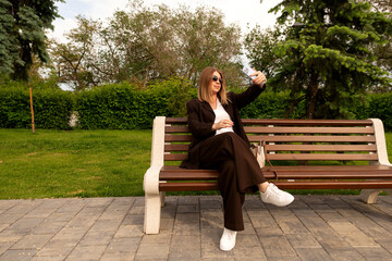 Fototapeta premium Young stylish woman takes selfie while seated on bench outdoors.