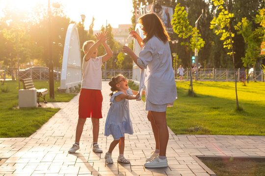 Golden-hour scene mother blowing soap bubbles while her laughing son and daughter jump and reach on paved park path near white bridge, enjoying carefree summer play