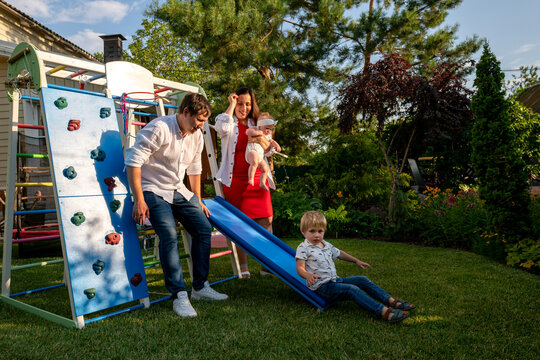 Little boy lands at the end of a blue slide while his parents, holding a baby, supervise beside a colorful backyard playset under warm evening light - Powered by Adobe