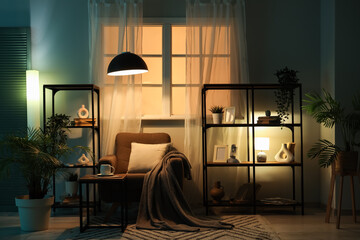 Interior of dark living room with armchair, shelf units and glowing lamps at night