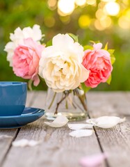 Obraz premium Roses in a glass vase with a blue cup and saucer on a wooden table outdoors in sunlight