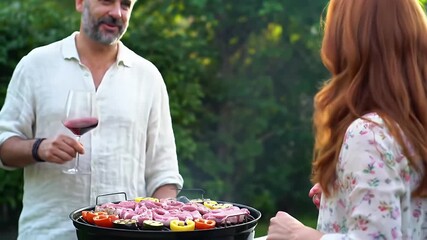 Smiling Couple Grilling Meat and Vegetables Outdoors on a Sunny Summer Day Bright Backyard Setting Lush Green Foliage Red Wine Glass Eye Level View - Powered by Adobe