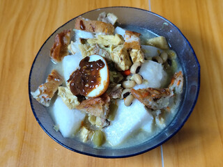 A bowl of vegetable rice cake. Lontong vegetable with coconut milk sauce and accompaniments of Indonesian tofu, beef and petis eggs, served on a wooden table.