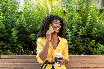 African-American woman in yellow blazer smiling while talking on smartphone and holding coffee cup,...