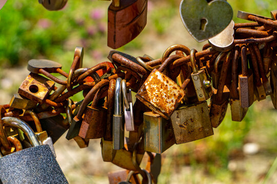 Rusty old love padlocks in various shapes and sizes, attached to a thick metal chain against a blurred green background.