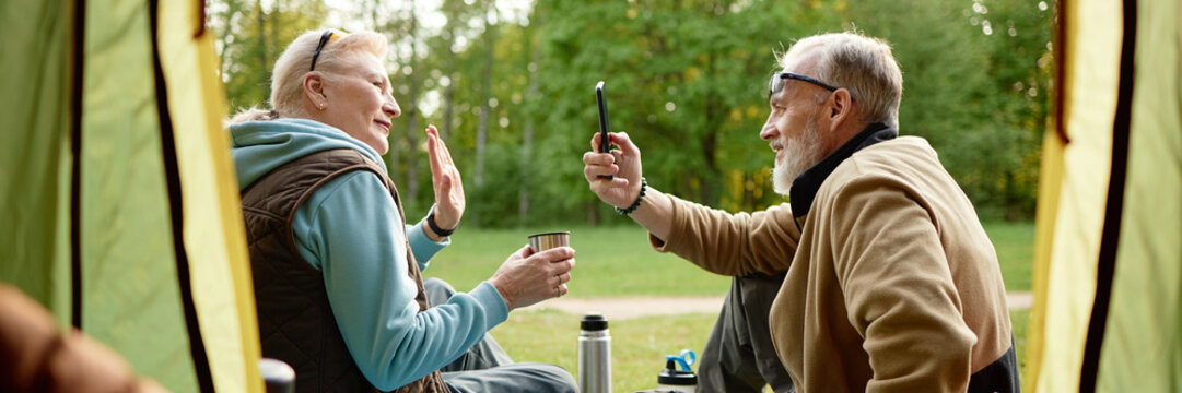 Header of mature Caucasian woman waving while senior Caucasian man holding smartphone taking photos outdoors, both sitting inside tent with forest background, enjoying camping activity - Powered by Adobe